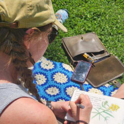 A person wearing sunglasses and a baseball cap sits in the sun and sketches a flower in a notebook