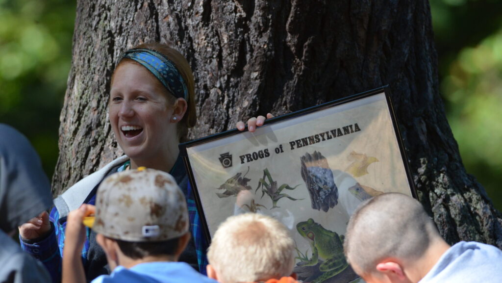 An outdoor educator holds a poster showing illustrations of the frog species that live in Pennsylvania