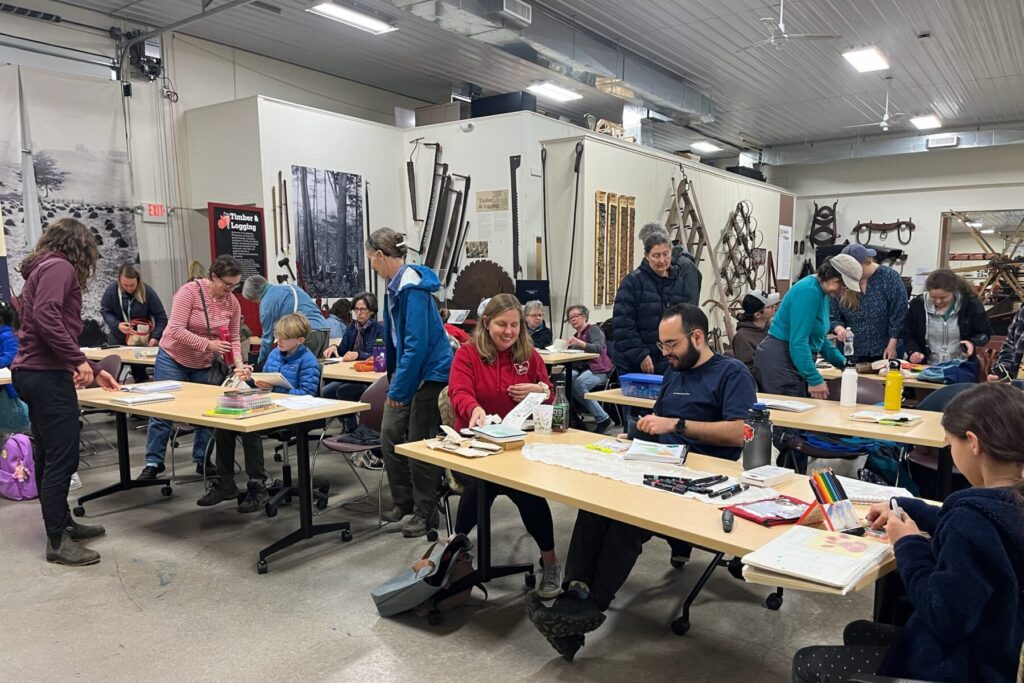 People sit at tables with notebooks and art supplies.