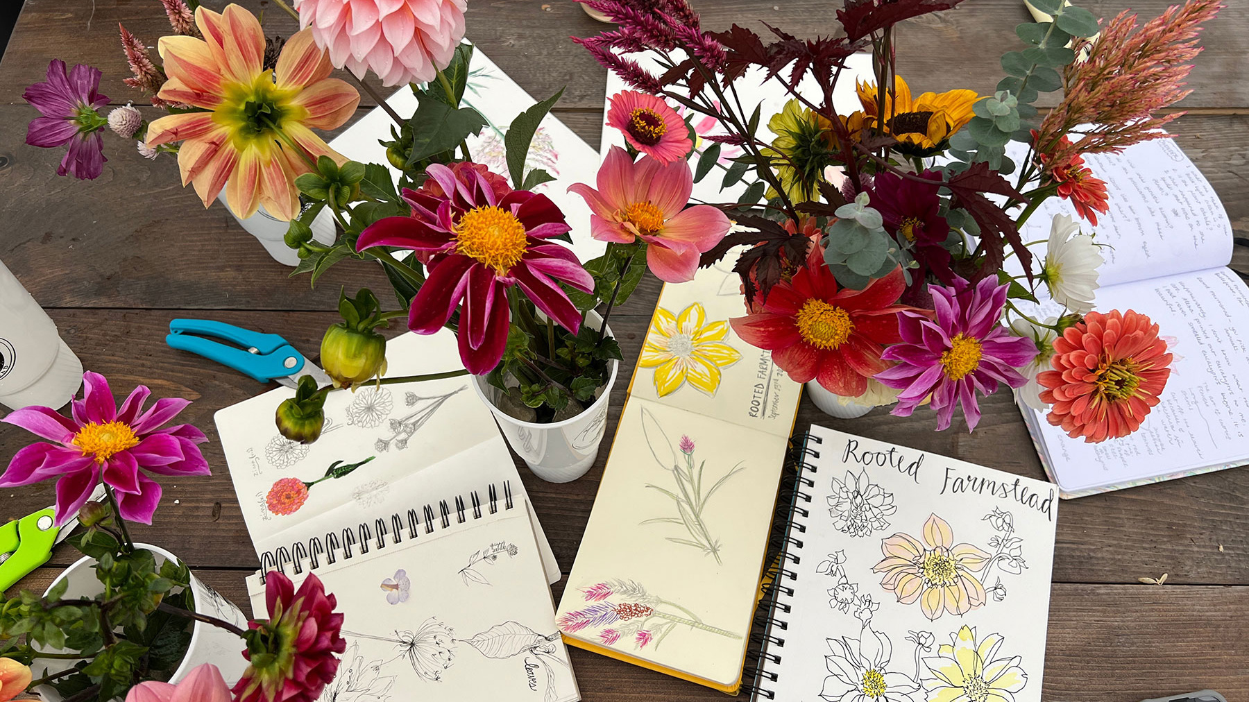 Flower arrangements and journals showing sketches of flowers are spread across a wooden table